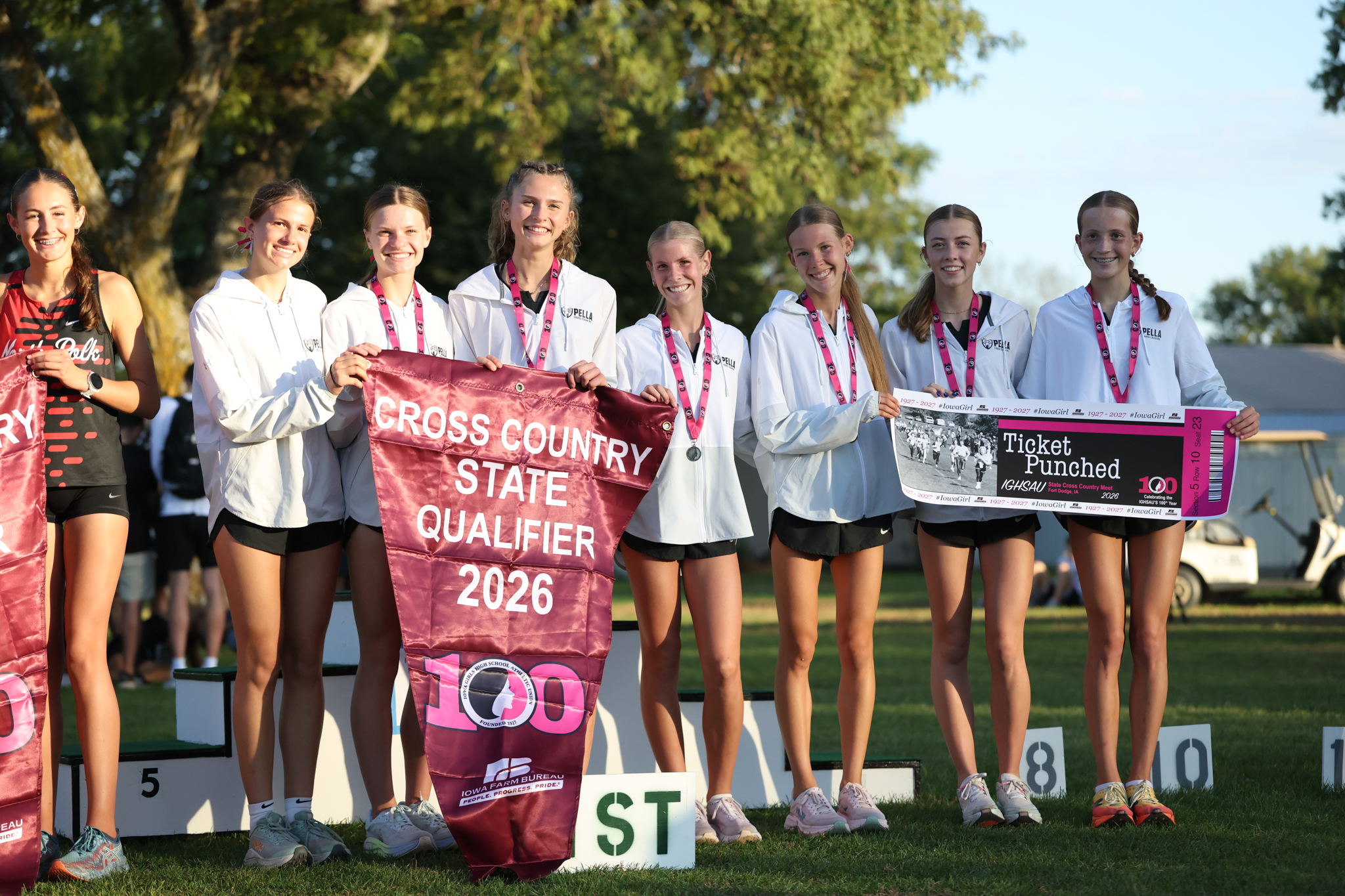 Seven Pella girls cross country runners smile with medals, holding a 2026 State Qualifier banner and a "Ticket Punched" sign.